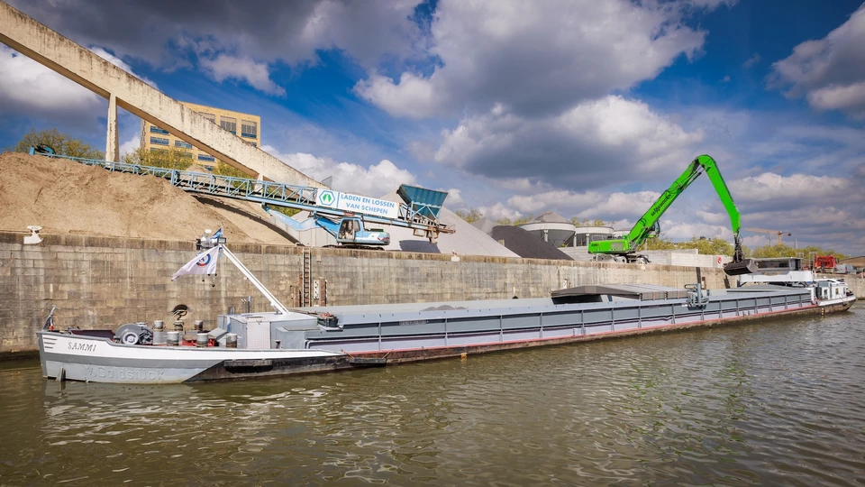 Een binnenschip ligt aangemeerd aan een kaai van de Haven van Brussel terwijl een kraan bulkgoederen van het schip lost. Op de achtergrond zijn haveninstallaties en opslagplaatsen te zien die de laad- en losactiviteiten via de waterweg illustreren.