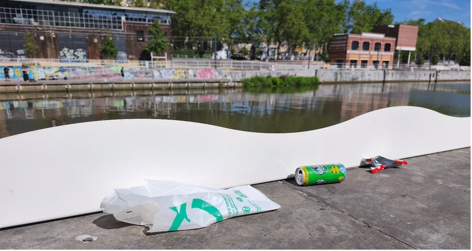 Zicht op een golvende witte afvalplint langs een kade van het Brussels kanaal. Een plastic zak, een blikje en enkele verpakkingen worden tegen de plint tegengehouden, wat illustreert hoe ze voorkomt dat afval in het kanaal terechtkomt.